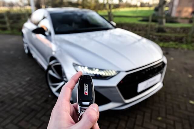 A hand holding a car key fob in the foreground with a grey coupe in the background