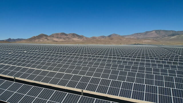 A wide field of solar panels in the foreground, with desert mountains and blue sky in the background