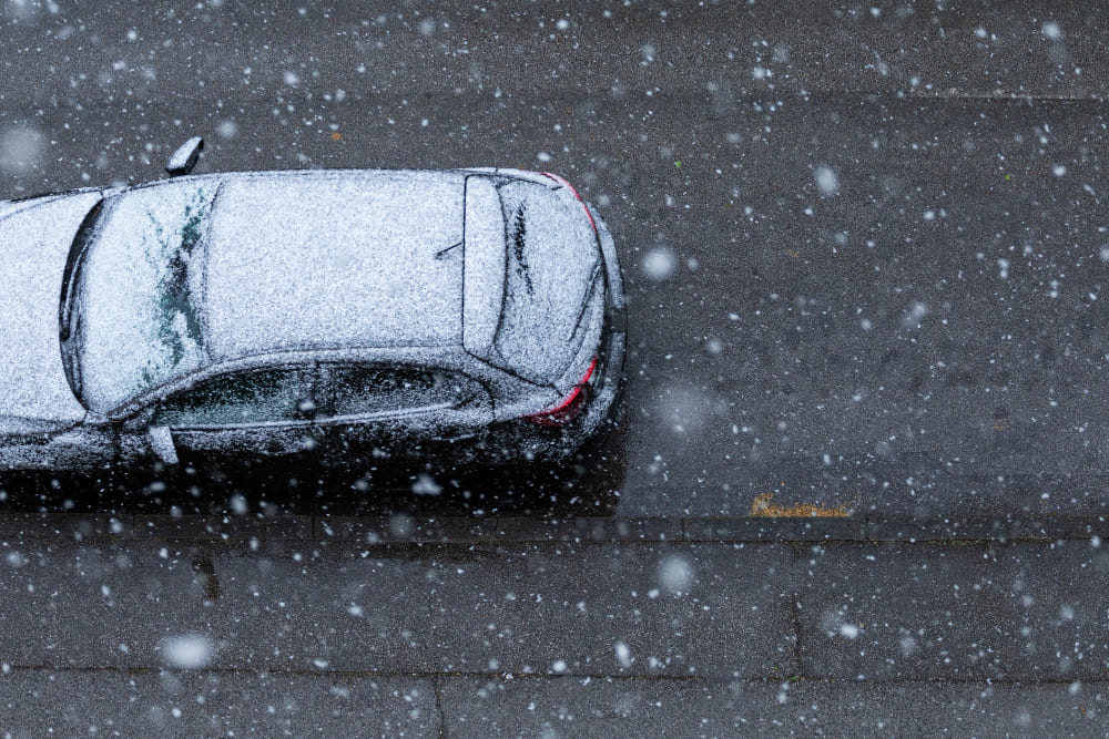 Overhead view of a car blanketed in snow parked next to the sidewalk, with snowfall in progress