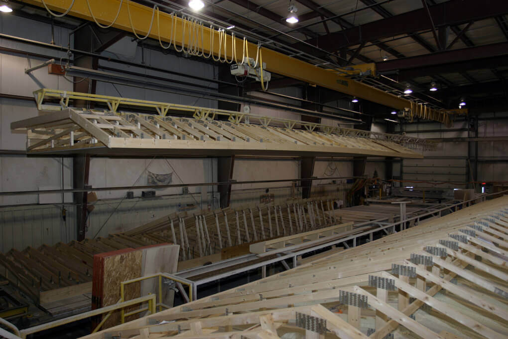 The interior of a modular home factory, with an unfinished roof suspended above a long line of trusses