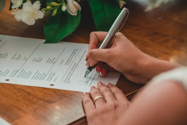 A hand holding a grey pen signs the bottom of a form. A white flower is visible in the background.