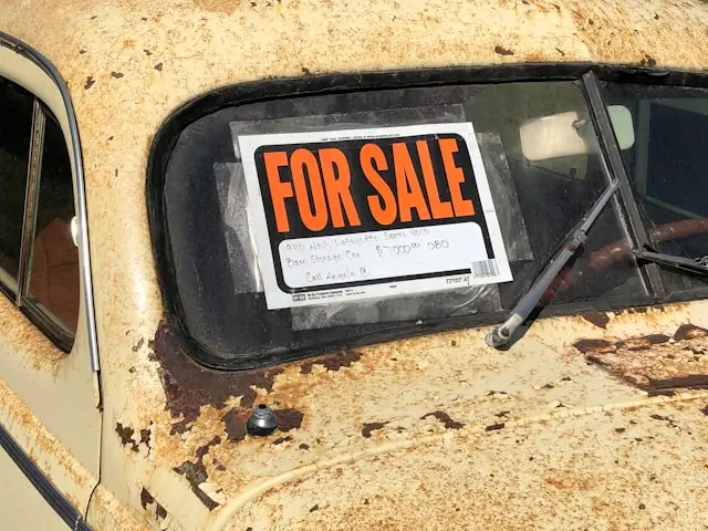 An orange 'for sale' sign in the rear window of a very old, rusty used car.