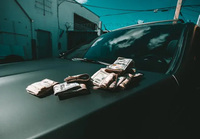 Used car scams and how to avoid them Bundles of cash piled on a matte black hood of a car outside a razor-wire-fenced building